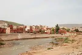 A river runs across the image from left to right with a town in the background, behind a concrete flood defence. The foreground shows a stony, sparsely vegetated river bank.