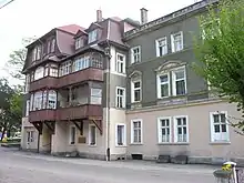 Bâtiment de quatre étages. Les murs du premier sont de couleur beige, et le reste est gris. Le toit est brun. Le bâtiment a deux balcons en bois sur sa face.