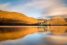 Un lac très calme. Au dessus, des collines ocres brillamment éclairées par le soleil levant ; au débouché d'une vallée, au bord du lac, un chateau fort.