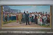 Peinture murale montrant Kim Jong-Il au centre, entouré par une foule compacte de personnes, l'université est visible dansle fond.