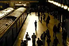 Un quai de gare vue en légère plongée, avec deux trains côte à côte à l'arrêt. La lumière du soir est vive et rasante et une silhouette en contre-jour se distingue au bord du quai, parmi la foule des passagers