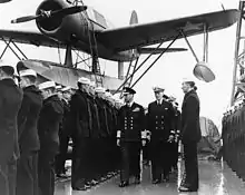 Photographie de plusieurs officiers passant en revue des marins. Un hydravion se trouve à l’arrière-plan.