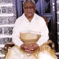 Photo en couleur d'un homme âgé ceint d'une natte traditionnelle (ta'ovala) et vêtu d'une chemise blanche, assis sur un trône en bois, devant une grande porte en bois et un mur en pierres apparentes.