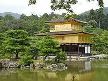 Photo couleur d'un édifice religieux carré à trois étages et aux façades dorées, se reflétant dans l'eau d'un étang, au premier plan, sur fond de forêt verdoyante.