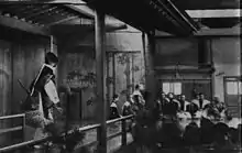 Premier plan à gauche : l'acteur s'avance sur le pont ; à droite, les spectateurs vus de dos. Au fond, musiciens accroupis.