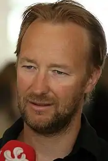 Portrait d'un homme aux cheveux et à la barbe blonds et courts, parlant dans un micro.