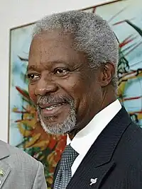 Photo de Kofi Annan, cheveux courts et blancs, moustache et barbe courte sur le menton, en costume noir, cravate, et chemise blanche, arborant un léger sourire.