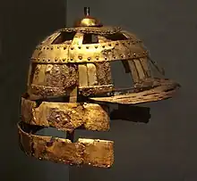 Photo couleur d'un casque de soldat fait de lames de fer recouvertes de cuivre, sur fond sombre.