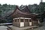 Petit bâtiment en bois avec des murs blancs et le toit en croupe et à pignon relié à une autre structure en bois à l'arrière.