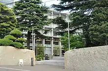 Photo couleur de l'entrée d'un campus universitaire. Deux murs d'enceinte beiges, à droite et à gauche, délimite un ouverture sur un escalier en pierre menant à l'entrée d'un bâtiment blanc à la façade vitrée, partiellement cachée par des arbres au feuillage vert.