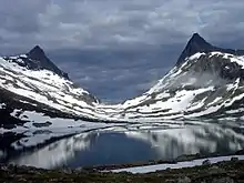 Pics rocheux de part et d'autres d'une vallée en U, avec un petit lac au premier plan et des plaques de neige un peu partout.