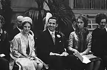 Photographie montrant trois personnes assises : un homme vêtu d'un costume trois pièces et deux femmes, l'une portant un manteau clair et un chapeau turban et l'autre un manteau brodé, une toque et une écharpe en fourrure