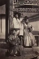 Photographie ancienne d'une groupe de trois hommes en costume taditionnel. L'un fume une longue pipe, et il sont réunis dans l'angle d'un bâtiment ouvrant vers l'extérieur.