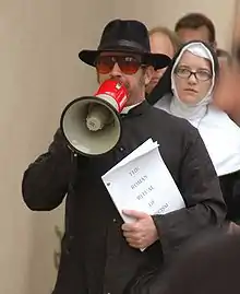 Photo d'un homme avec un chapeau noir et des lunettes de soleil qui parle dans un mégaphone.
