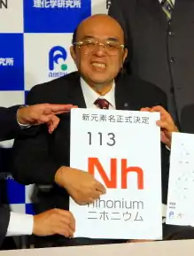 Photographie d'un homme tenant une affiche portant l'inscription « Nihonium » associé au symbole et au numéro atomique de l'élément.