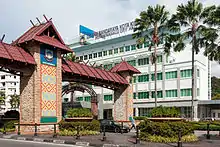 Photographie. Au premier plan se trouve l'entrée de l'hôtel de ville de Kota Kinabalu qui est un portique constitué de deux piliers sur lesquels se trouvent les armoiries de la ville. Au second plan se trouve l'immeuble faisant office d'hôtel de ville.