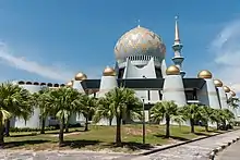 Photographie d'une mosquée blanche et dorée, de son grand dôme, de son haut minaret et des quelques palmiers qui l'entourent.
