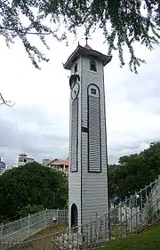 Photographie d'une grande tour blanche surmontée d'un toit pyramidal. Sur sa façade gauche se trouve une porte d'entrée en bas et une horloge en haut. Au sommet du toit se trouve une girouette.
