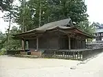 Bâtiment en bois avec un toit en croupe et à pignon et auvent. Une plus courte allée est fixée sur le côté droit.