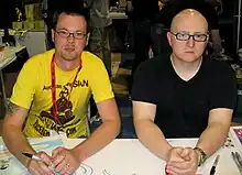 Deux hommes en t-shirt assis derrière une table regardent la caméra.