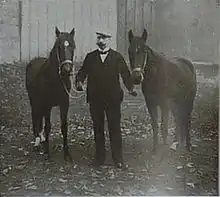 Photo en noir et blanc représentant un homme en costume tenant en main de part et d'autre deux chevaux en licol.