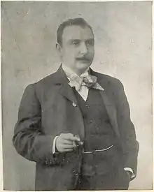 Photographie d'un homme moustachu, en costume, nœud papillon et cigare à la main droite.