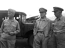 Photographie en noir et blanc de trois hommes vêtus de costumes militaires clairs et se tenant devant un véhicule militaire.