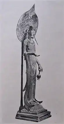 Vue de trois-quarts d'une statue grande et très mince portant un vase avec deux doigts de sa main gauche. Son bras droit est courbé avec la paume de sa main droite vers le haut. Une auréole sur un mât est vue derrière la statue.