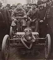 Photo de face de deux hommes assis dans une voiture rudimentaire et portant le numéro de course 20.