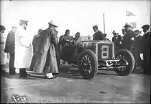 Photo de deux hommes en tenue de pilote dans une voiture prête à prendre le départ d'une course devant des officiels.