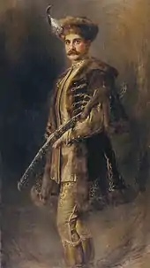 Peinture d'un homme en pied habillé d'un costume hongrois traditionnel recouvert d'un manteau de fourrure, le visage grave orné de moustaches, il presse contre son corps de son bras gauche une épée finement ciselée.