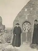 Deux personnes en soutane noire, celui à gauche portant une canne et un chapeau, se tiennent près d'une chapelle en pierre (on voit une porte en bois)