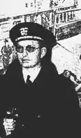 L. Ron Hubbard avec lunettes et blouson militaire devant un navire militaire.