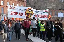 Photo couleur de manifestants défilant dans une rue et portant une banderole blanche sur laquelle on peut lire « Stop au nucléaire ».