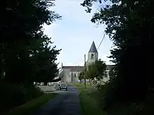 Une petite route bordée d'arbres avec au fond une église de pierre couverte de tuiles au clocher conique ardoisé. Un panneau de limitation de vitesse à 30 km/h à droite.