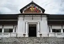 Photo contemporaine de la façade d'un palais aux murs blancs, dont le porche est orné d'un bas-relief doré et rouge représentant deux éléphants.