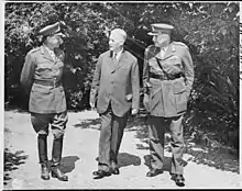 Photographie en noir et blanc de trois hommes debout et discutant, celui du milieu habillé en civil, les deux autres en uniforme militaire.