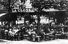 Vue en noir et blanc d'une terrasse de café bondée de consommateurs assis, au début du XXe siècle.