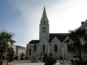 Photographie d'une place ornée de palmiers en bacs. Au centre, statue de bronze sur un haut piédestal. Au second plan une église : le chœur coté gauche, le clocher au centre (à la croisée des transepts).