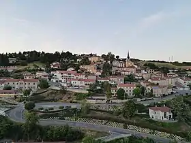 Une photographie du bourg prise de façon à ce que l'ensemble du centre de la commune, pourtant relativement escarpé, soit visible. On y voit une route qui serpente au premier plan, bordée de verdure et surplombée par un grand nombre de maisons individuelles, souvent aux murs beiges et aux toits de briques rouges. Le bâtiment contenant la mairie et l'école du village, sur la gauche, ressort particulièrement. En fond, des bois couvrent la ligne d'horizon.