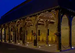 Photographie nocturne en couleurs d'une halle ouverte sur trois côtés. Des piliers en bois soutiennent un étage couvert d'ardoises et de tuiles.