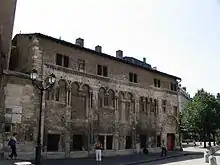 Photographie représentant un bâtiment doté de voûtes romanes, situé en ville devant une place.