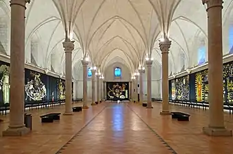  Photographie d'une salle voutée ogives où sont accrochées des tapisseries à dominante noir et or.