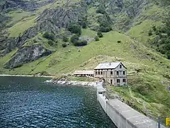 Photographie couleur d'un barrage en béton, retenant un lac. À l'arrière-plan, un refuge et des montagnes.