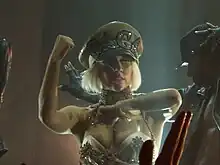 Une femme blonde vêtue d'un bustier khaki et d'un chapeau d'amiral sur lequel est écrit GAGA est visible. Elle regarde vers la droite de l'image. Sa main droite est positionnée devant son coup et sa main gauche est placée verticalement prêt de son visage. Une silhouette d'un homme dansant est visible.