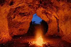 Photographie en couleurs d'un feu consumant des bûches en bois, sa lumière rougeoyante se réfléchissant sur les parois d'une grotte.