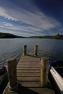 Un quai en bois s'avance dans un lac. À l'arrière-plan, des nuages couvrent partiellement le ciel bleu.