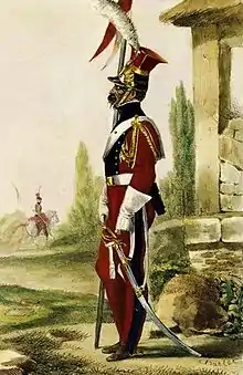 Un soldat, de profil, vêtu d'un uniforme rouge et bleu, tenant une lance dans sa main droite, à côté d'un bâtiment en pierre.