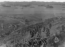Photographie prise depuis une colline donnant sur une large baie où se trouvent de nombreux navires de transport. Au premier plan, une colonne de soldats en armes marche sur un sentier dans un paysage aride et pelé.