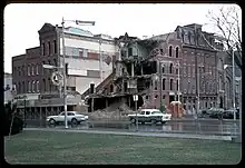 Photo prise en janvier 1966 du centre-ville de Norwich, où un grand magasin à la jonction des rues Main (NY 319) et Broad (NY 12) est en train d’être démoli. Le premier marqueur qui permet de savoir qu'on est sur la route NY 319, en direction ouest, se trouve en bas à droite. On peut y apercevoir des voitures, typiques des années 60, rouler sur NY 319.